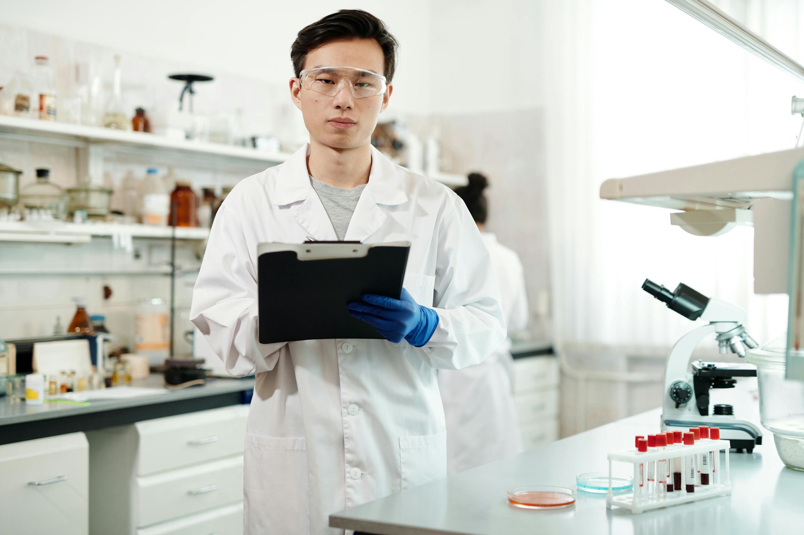 A young male scientist wearing a lab coat and safety glasses analyzes data in a modern laboratory setting.